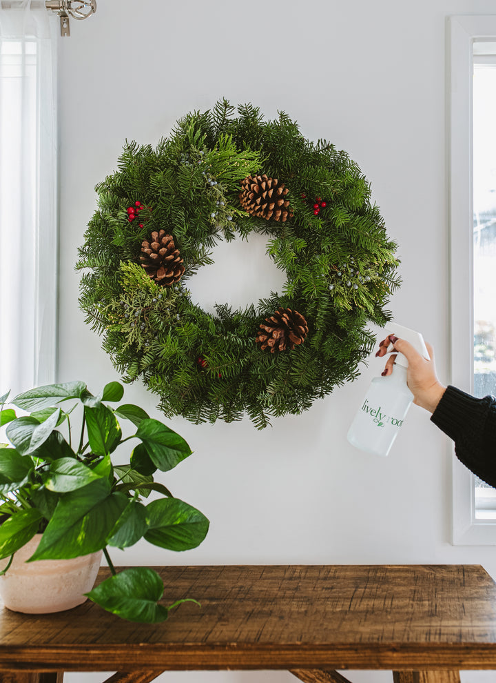 Lively Root white plant mister spray bottle with minimalist branding being used to mist evergreen holiday wreath with pinecones and berries, perfect for plant care and seasonal home décor maintenance.