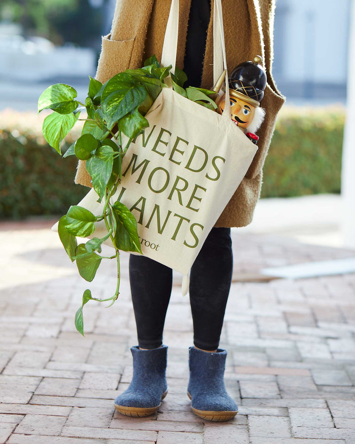 "Needs More Plants" canvas tote bag styled in outdoor lifestyle setting with flowering houseplant and holiday décor, perfect for plant-themed gifts, garden accessories, and eco-conscious home décor storage.