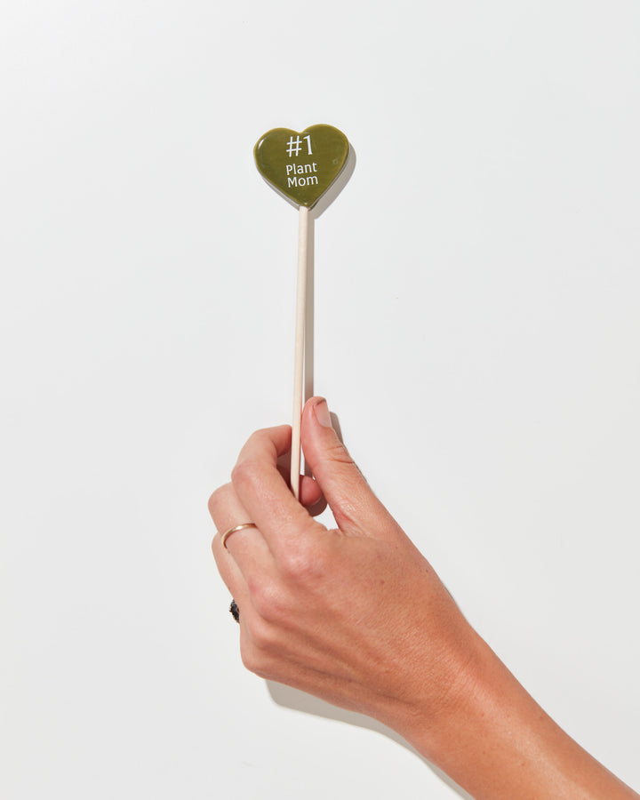 Olive green heart-shaped plant pick with "#1 Plant Mom" engraved text and natural wooden dowel stem, perfect decorative plant marker for houseplant lovers and plant parent gift.